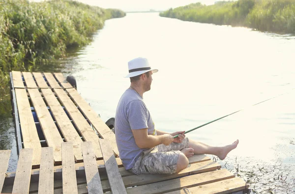 Side view from above of barefoot man in casual wear and hat sitting on wooden bridge and fishing with rod on tranquil river among green plans