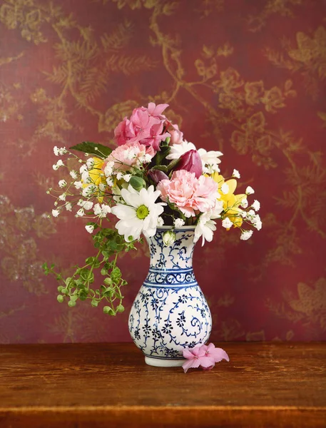 白色花瓶上的一束簇五彩缤纷的花朵 镶嵌着蓝色的装饰品 贴在木制桌子上 贴着金黄色图案的红葡萄酒墙纸 静谧的生活 — 图库照片