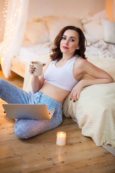 Optimistic lady with modern laptop and mug of hot beverage looking at camera while sitting near bed and candle in cozy bedroom