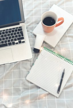 Dizüstü bilgisayar ve kalemin yanına dizüstü bilgisayar ve bir fincan kahve ve kitap ve kozmetik tüple süslenmiş gri kareli kumaşla süslenmiş, açık renk çelenkle süslenmiş.  