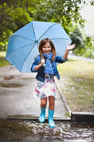 公園で遊んでいる間 青い傘が笑っているとカメラで笑みを浮かべて水たまりにジャンプしているカジュアルな服やゴムブーツで遊び心のある就学前の子供 — ストック写真