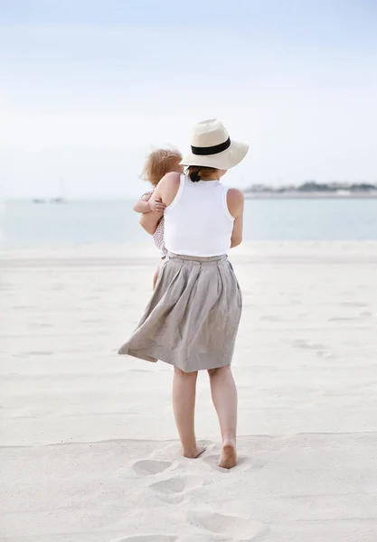 夏の日に海辺の白い砂の上に立っている間 赤ちゃんを保持灰色のスカートと帽子と白いタンクの上に身を包んだ若い母親のバックビュー — ストック写真