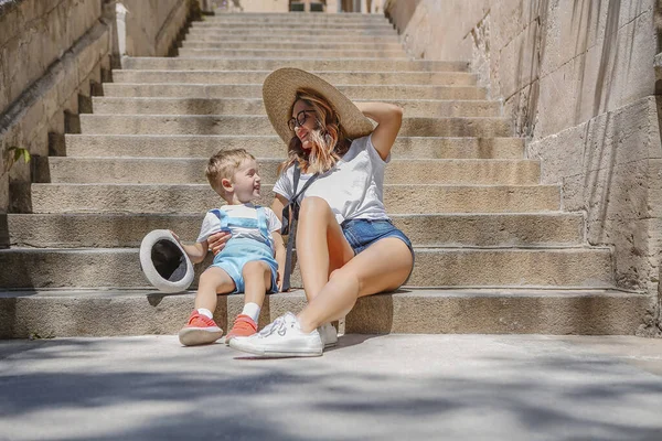 街の通りで夏の日にみすぼらしい階段に座っている間 全身の幸せなスタイリッシュな女性と陽気な幼児はお互いを見て笑っています — ストック写真