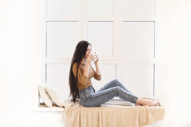 Sıradan giyinmiş genç bir kadının pencere pervazında oturup evde dinlenirken çay içişini yan görüyorum.