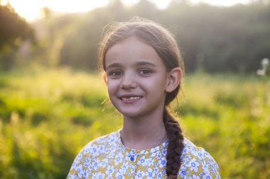 Renkli atkuyruklu neşeli küçük kız yaz akşamları yeşil tarlada dikilirken gülümseyip kameraya bakıyor.