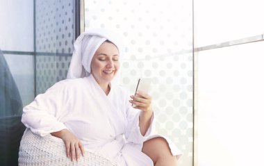 Yeni akıllı telefonunu kullanırken balkonda bornoz giyen genç bir kadın. 