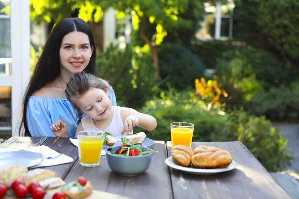 幸せな若い女性でカジュアル摩耗に座ってテーブルと小さな娘を保持しながら 健康的な夕食と新鮮なサラダとパンとオレンジジュースで庭で晴れた夏の夜 — ストック写真