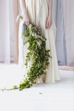 Çıplak ayaklı, zarif, beyaz gelinlikli, büyük bir buket beyaz çiçekli, yeşil yapraklı, şeffaf pastel perdelerin arasında duran bir kadın.