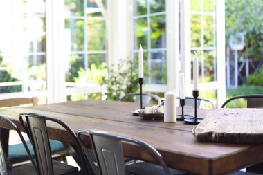 Güneşli bir yaz gününde bahçedeki cam terasta, ahşap masaya yerleştirilmiş beyaz mumlar ve tabaklar.