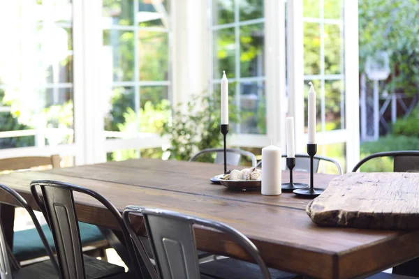Güneşli bir yaz gününde bahçedeki cam terasta, ahşap masaya yerleştirilmiş beyaz mumlar ve tabaklar.