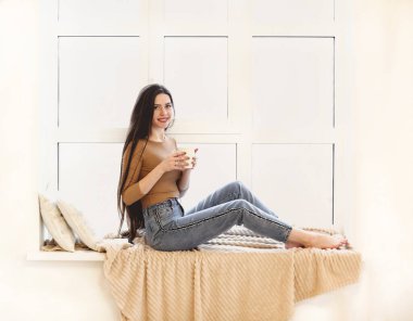 Sıradan giyinmiş genç bir kadının pencere pervazında oturup evde dinlenirken çay içişini yan görüyorum.