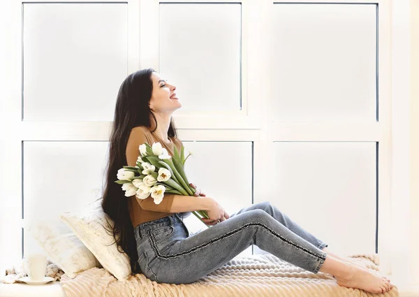 白いチューリップの花束を保持し 自宅で居心地の良い部屋の窓辺に座っている間笑みを浮かべてカジュアルな服装で幸せな若い女性の側面図 — ストック写真