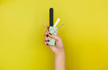 Parlak sarı arka planda çeşitli manikür aletleri gösteren tırnakları boyalı tanınmayan bir kadın.