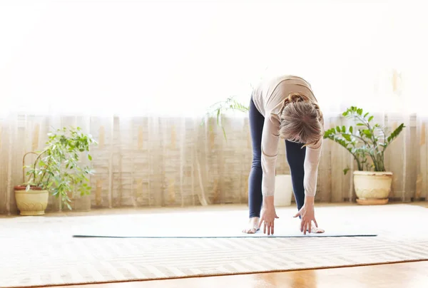 Full body adult female in casual clothes raising arms and stretching body while doing yoga on floor at home