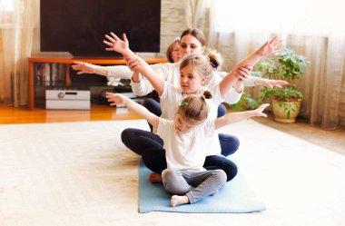 Yetişkin kadınlar ve küçük kızlar evde yoga yaparken yerde oturup kollarını uzatırlar.