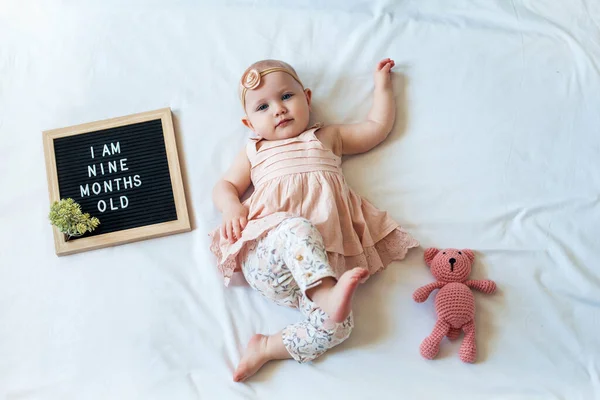 9 Nove mesi bambina sdraiata su sfondo bianco con cartolina e orsacchiotto. Composizione laica piatta . — Foto Stock