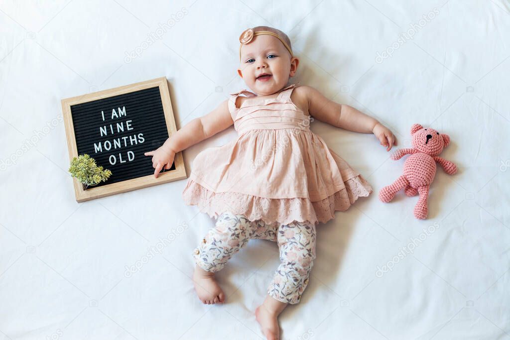 9 Nine months old baby girl laying down on white background with letter board and teddy bear. Flat lay composition.