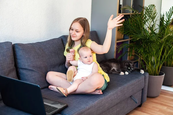 Щаслива дівчина з сестрою-немовлям дивиться на екран ноутбука і жестикулює привіт, маючи відеодзвінок . — стокове фото