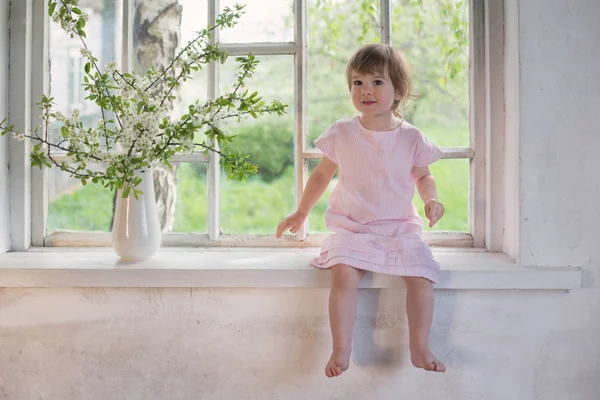 Belle petite fille sur le rebord de la vieille fenêtre avec des fleurs de printemps — Photo