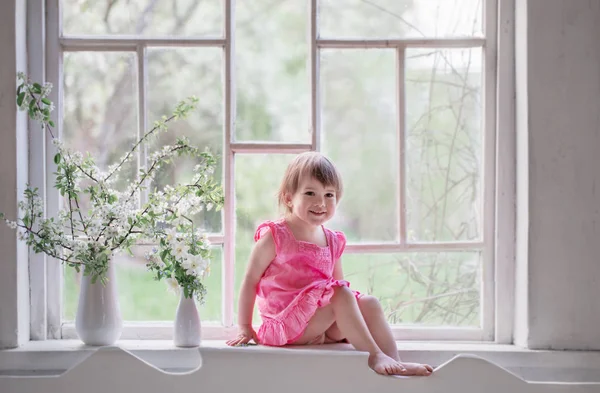 Beautiful little girl on old windowsill with spring flowers — Stock Photo, Image