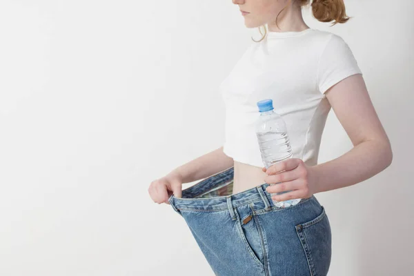 Chica con agua en botella tirando de sus pantalones vaqueros grandes y mostrando weig — Foto de Stock