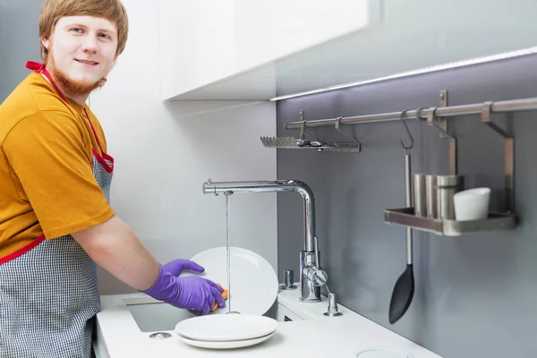 Молодой человек с рыжей бородой моет посуду на белой современной кухне — стоковое фото