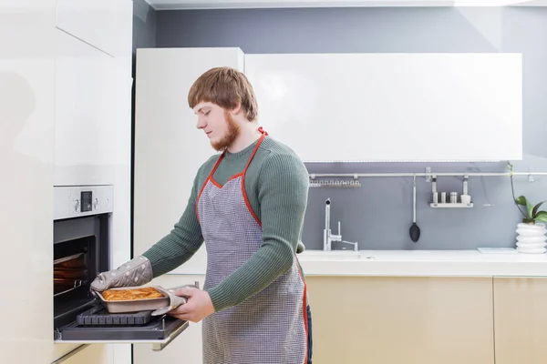 Ung man med rött skägg gör paj på vitt modernt kök — Stockfoto