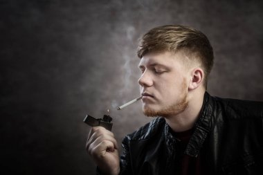 Deri ceketli genç adam siyah arka planda sigara içiyor.