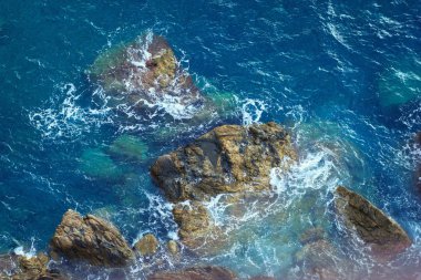 Kıyı şeridindeki taşlardaki deniz dalgaları üzerinde hava manzarası. Dağ kayaları sahilinde taşlı mavi deniz suyu.