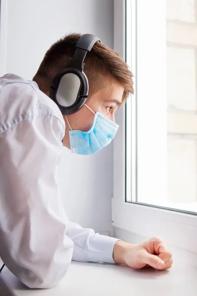 Young Man in Flu Mask and Headphones by the Window in the Room