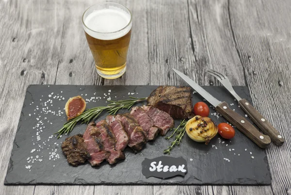 Filete de striploin a la parrilla en un plato de piedra con un vaso de cerveza con tenedor y cuchillo . —  Fotos de Stock