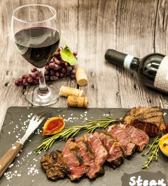 Primer plano del jugoso filete de ternera con una botella y una copa de vino tinto en un plato de piedra negra sobre una mesa de madera . —  Fotos de Stock