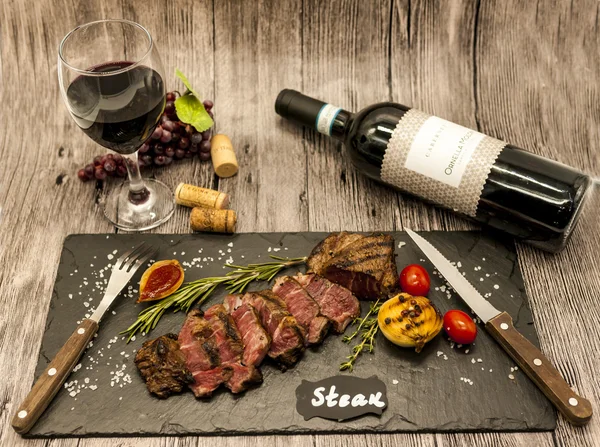 Nahaufnahme eines saftigen Rindersteaks mit einer Flasche und einem Glas Rotwein auf einem schwarzen Steinteller auf einem Holztisch. — Stockfoto