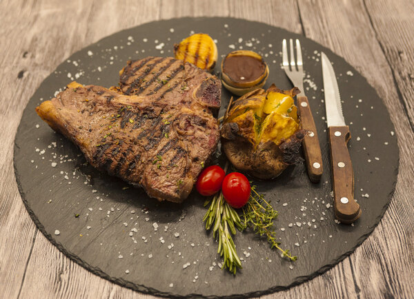 On wooden table background juicy beef steak medium rare on a stone baked potatoes and barbecue sauce and large sea salt with fork and knife.