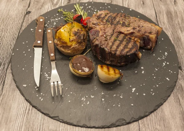 Sobre fondo de mesa de madera jugoso filete de res medio raro en una piedra al horno patatas y salsa barbacoa y sal marina grande con tenedor y cuchillo . —  Fotos de Stock