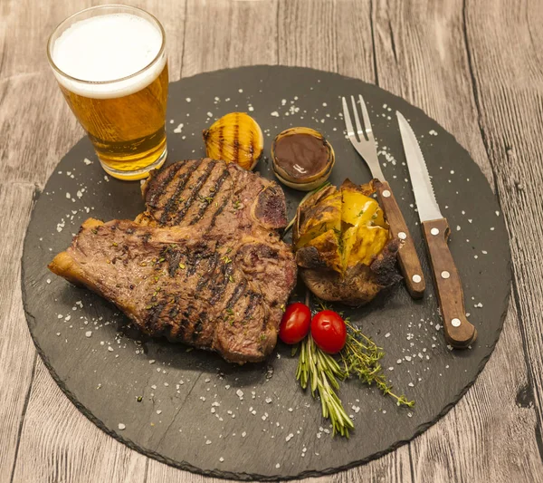 Su sfondo tavolo di legno succosa bistecca di manzo con un bicchiere di schiuma di birra leggera . — Foto Stock