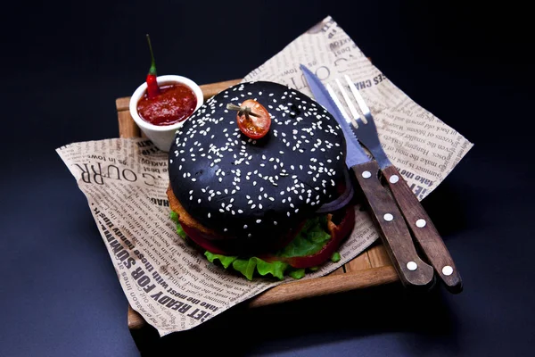 Saftige schwarze Fischburger. Burger mit Messergabel und Chilisoße auf der Zeitung — Stockfoto