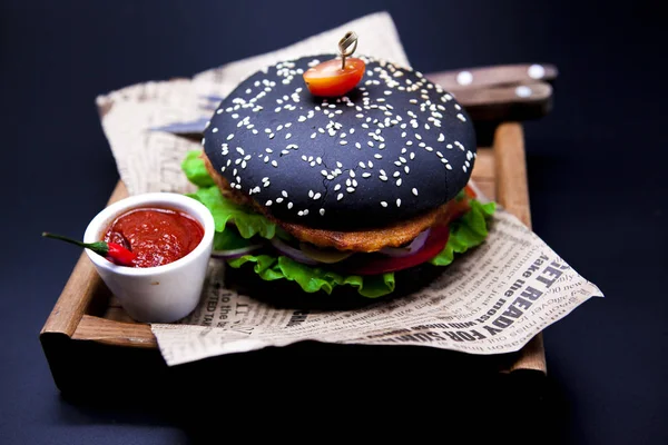 Saftige schwarze Fischburger. Burger mit Messergabel und Chilisoße auf der Zeitung — Stockfoto