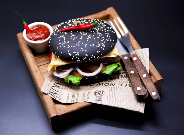 Šťavnatý černý burger. Burger s mramorovou hovězí, vidlice s nožem a chilli omáčka na noviny — Stock fotografie