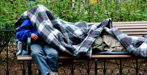 Homeless people sleeping under blankets on park benches outdoors.