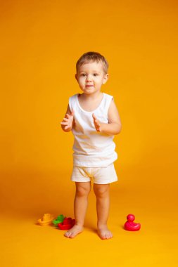 baby in a t-shirt and underwear stands next to his toys on a yel clipart