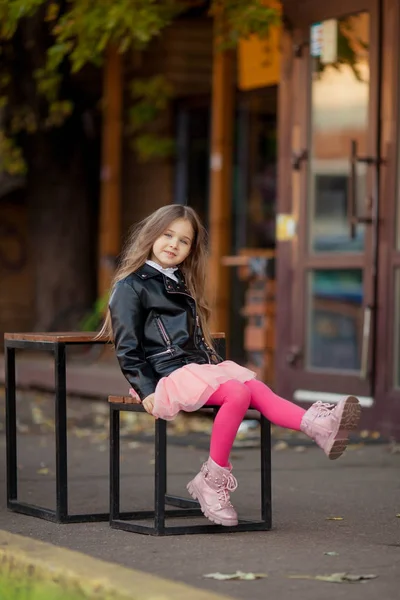 Menina de cabelos longos em uma jaqueta de couro acenando pernas em um banco — Fotografia de Stock