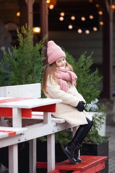 girl in winter clothes dangles legs sitting on a street decorated for Christmas