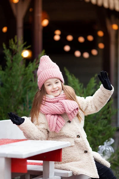 girl in winter clothes dangles legs sitting on a street decorated for Christmas
