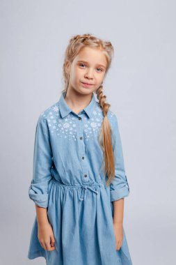 Uzun saçlı kız açık gri bir arka planda mavi bir elbiseyle duruyor..