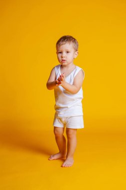 Beyaz tişörtlü ve külotlu bir çocuk turuncu-sarı arka planda duruyor.