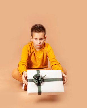 Yerde oturan çocuk elinde yeşil bir fiyonkla bir hediye kutusu tutuyor.