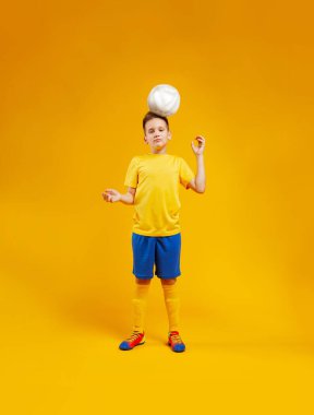Çocuk sarı arka planda, stüdyoda futbol oynuyor.