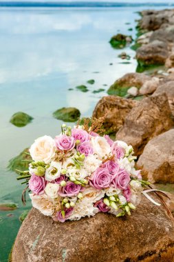 Mor ve beyaz gül renginde düğün buketi, deniz tarafından taşların üzerinde fotoğraflanmış.