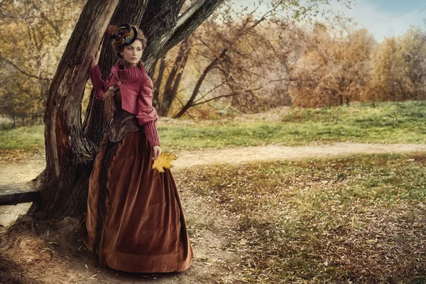 Woman in historical dress near the tree in autumn forest.